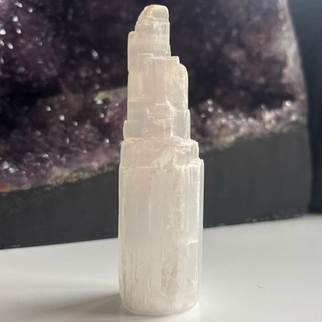 A polished white selenite crystal tower standing upright on a white surface indoors, showcasing a unique misshaped skyscraper profile, vertical fibrous lines, and a pearlescent shimmer styled in front of a dark amethyst geode for Crown Chakra alignment.