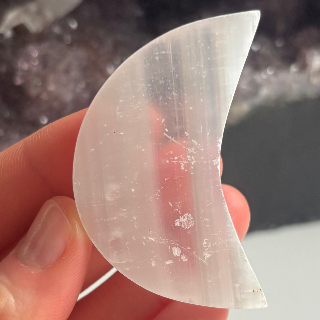 A close-up view of a polished white selenite half moon carving held between fingers indoors, showcasing its ice-like pearlescent shimmer, crescent silhouette, and natural fibrous layers for Crown Chakra alignment.