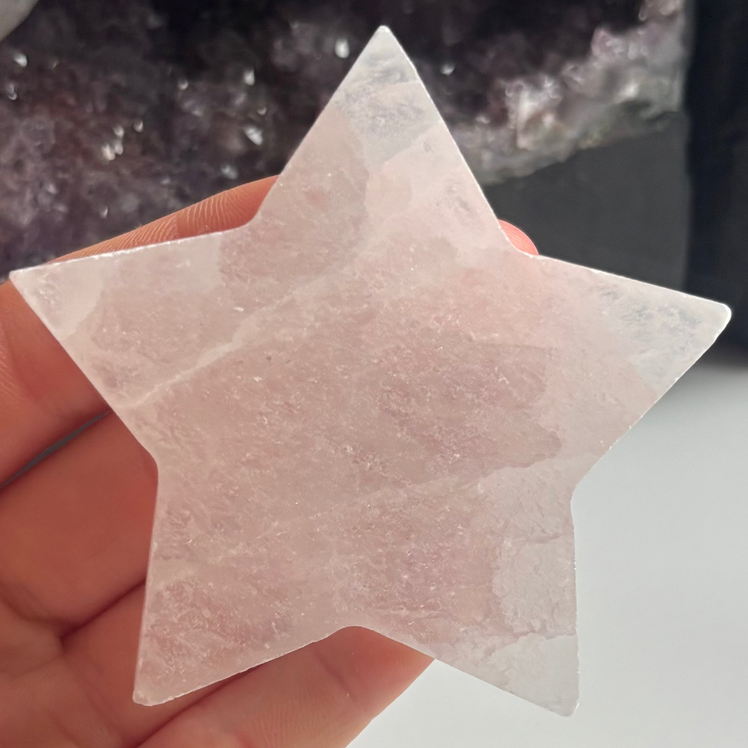 A close-up view of a large polished white selenite star carving held flat against a palm indoors, detailing its ice-like pearlescent shimmer, five-pointed cosmic shape, and soft fibrous layers for Crown Chakra alignment.