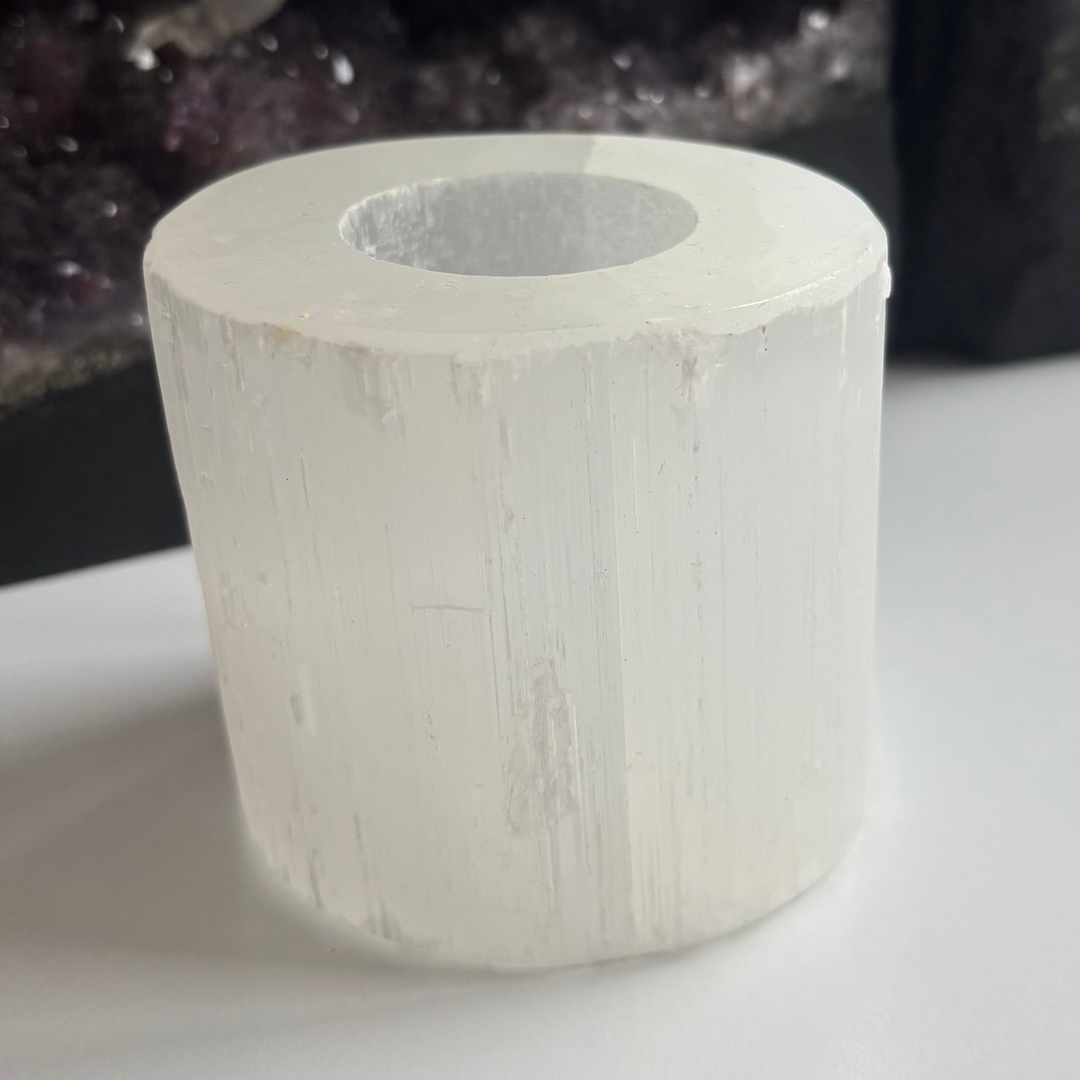 A raw white selenite tea-light candle holder standing upright on a white table layout, showcasing its heavy cylindrical tower shape, vertical mineral ridges, and natural ice-like texture in front of a dark amethyst geode for Crown Chakra alignment.
