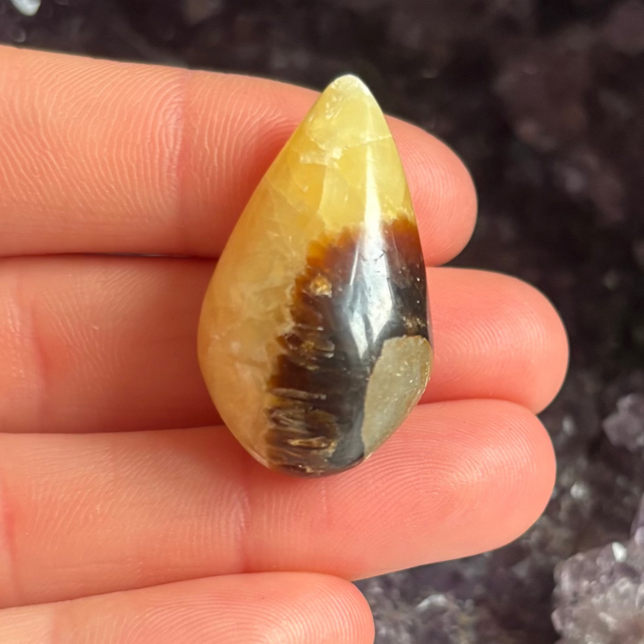 A close-up view of a polished septarian jasper tear drop carving held upright across fingers indoors, showcasing its unique split pattern of deep mud-brown lines and ancient golden tones against a dark amethyst geode for Root Chakra alignment.
