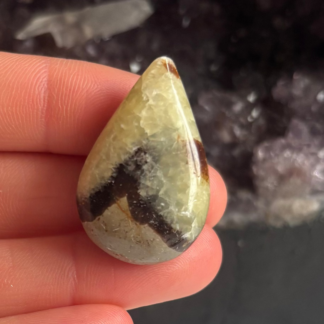 A close-up view of a polished septarian jasper tear drop carving held upright in fingers, showcasing a cream matrix split by bold, dark mud-brown zigzag veins against a blurred background of a deep purple amethyst geode for Root Chakra alignment.