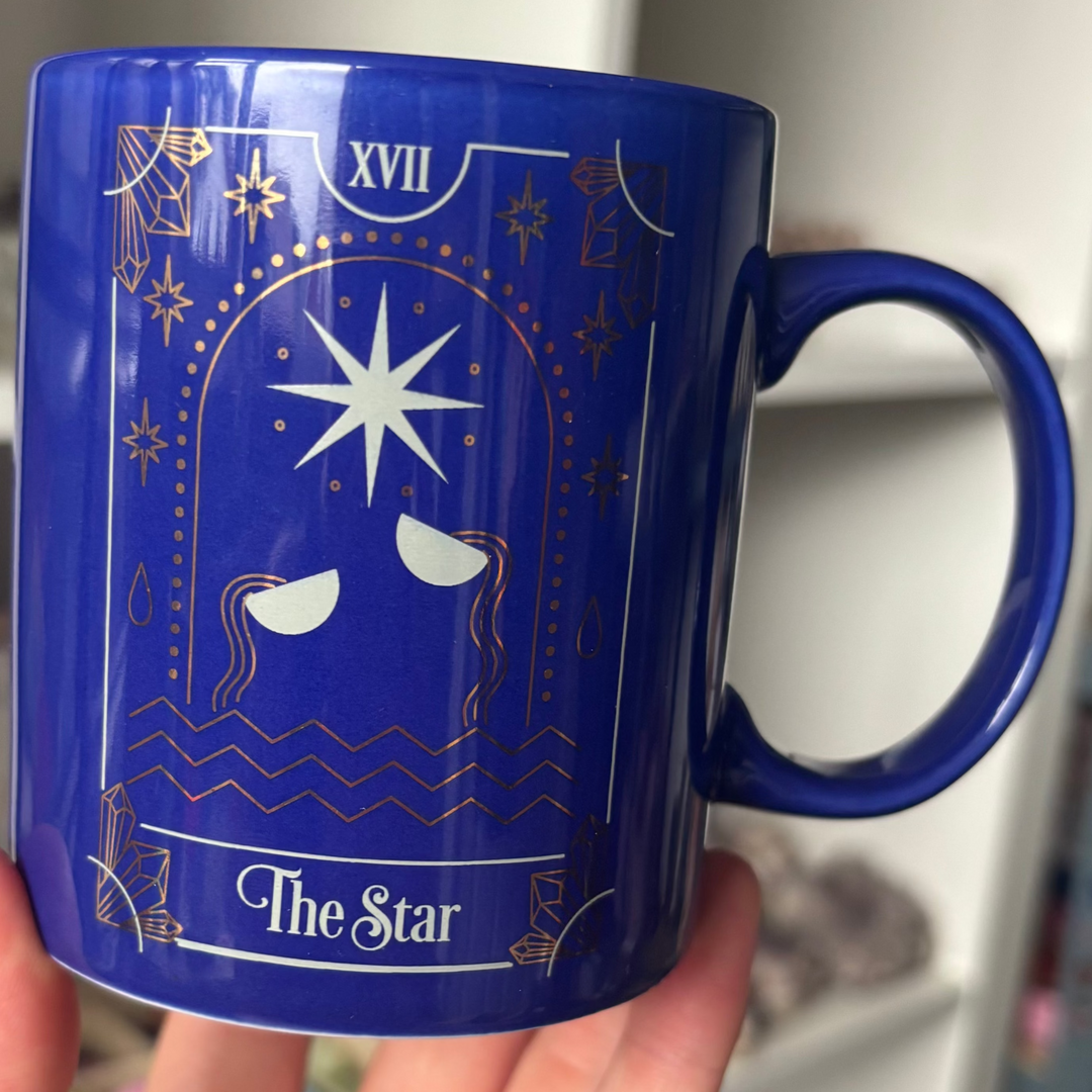A close-up view of the cobalt blue ceramic Star tarot card mug held gently by a hand. The glossy surface features beautiful gold and white celestial artwork detailing a prominent central star, perfectly matching the calm energy of the Third Eye and Crown chakras.