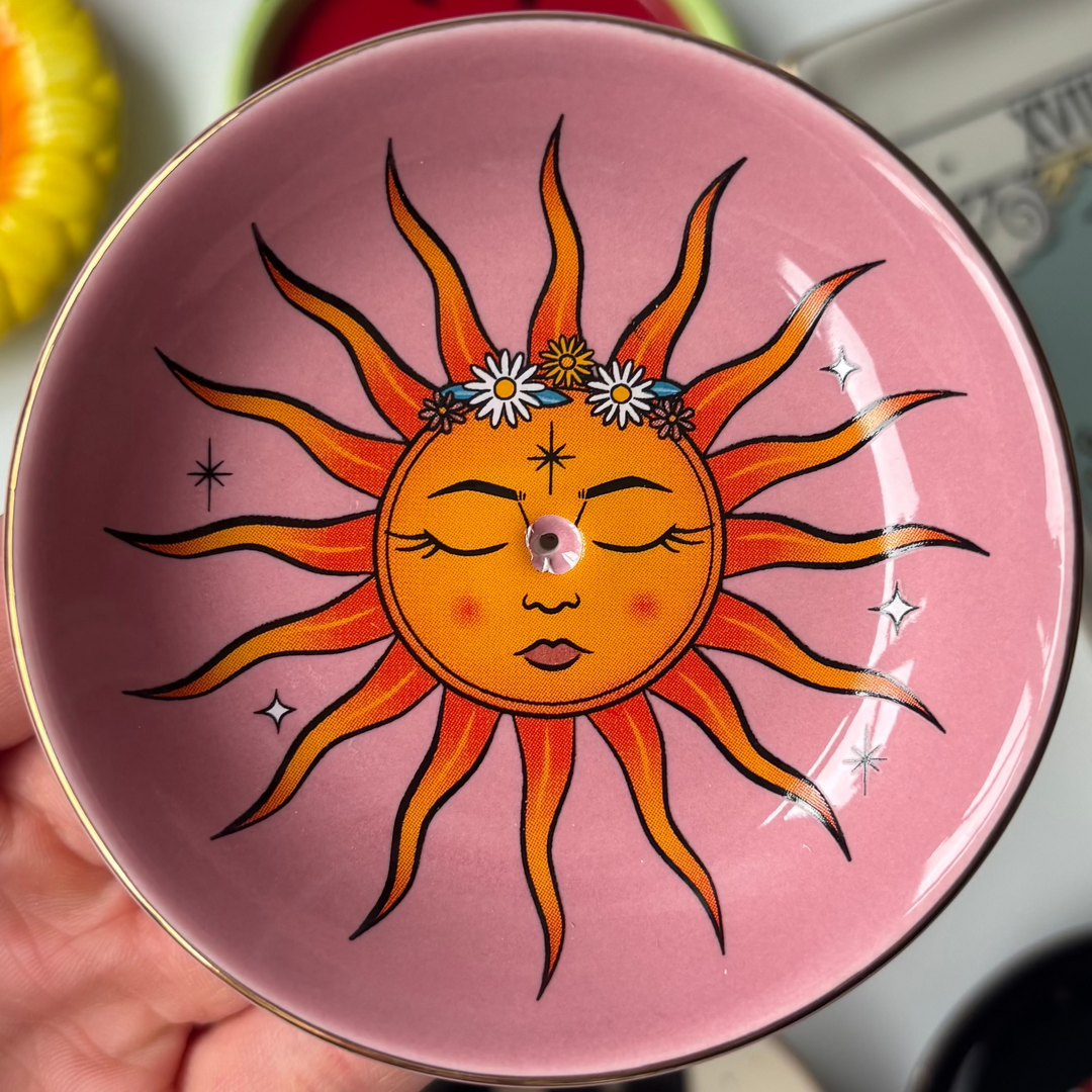 A close-up view of the round pink ceramic Sun celestial incense stick holder held gently in a hand. The glossy surface features a detailed solar design at its centre with a smiling face, vibrant orange sunbeams, and small stars to welcome bright vibes.