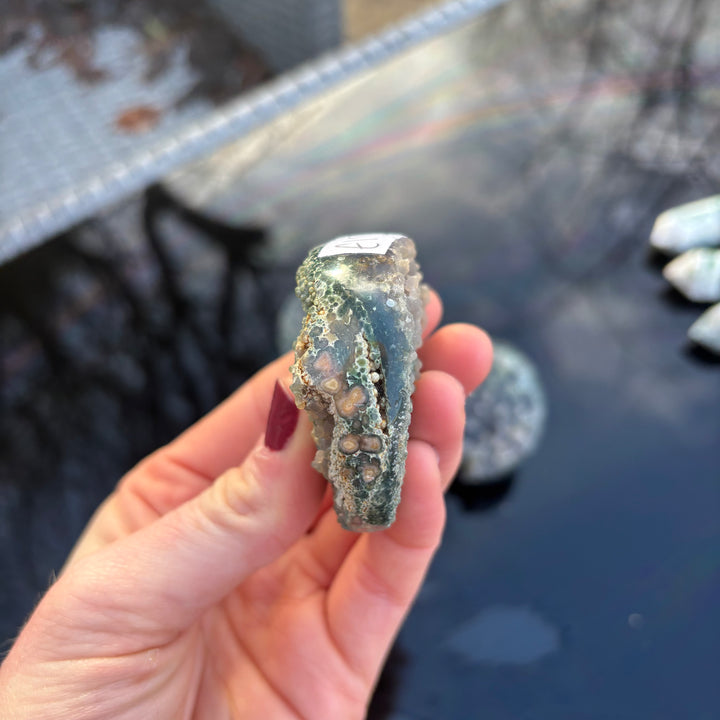 A side perspective view of the round Grape Agate crystal freeform held between fingers, displaying its thick, chunky edge and unique geological depth.