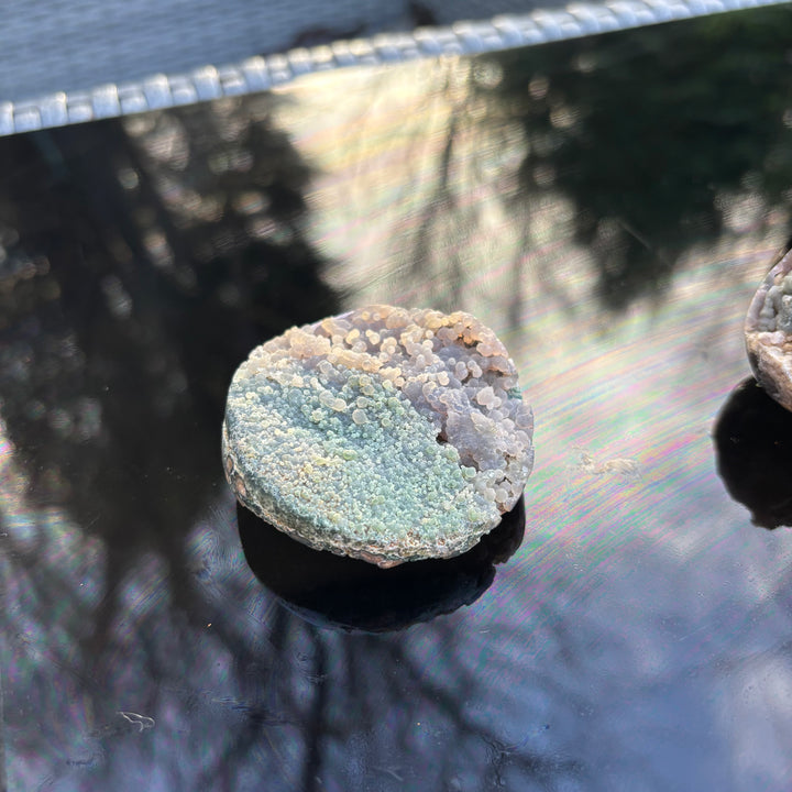 The polished circular Grape Agate gemstone block resting flat on a reflective black surface outdoors, ideal for third eye chakra alignment and whimsical home altar decor.