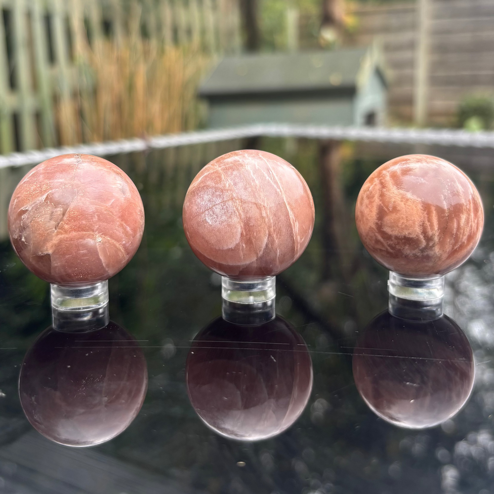 Three polished Peach Moonstone crystal orbs lined up side by side on their clear display stands outdoors, highlighting the natural shading variations and unique structural patterns across the collection.