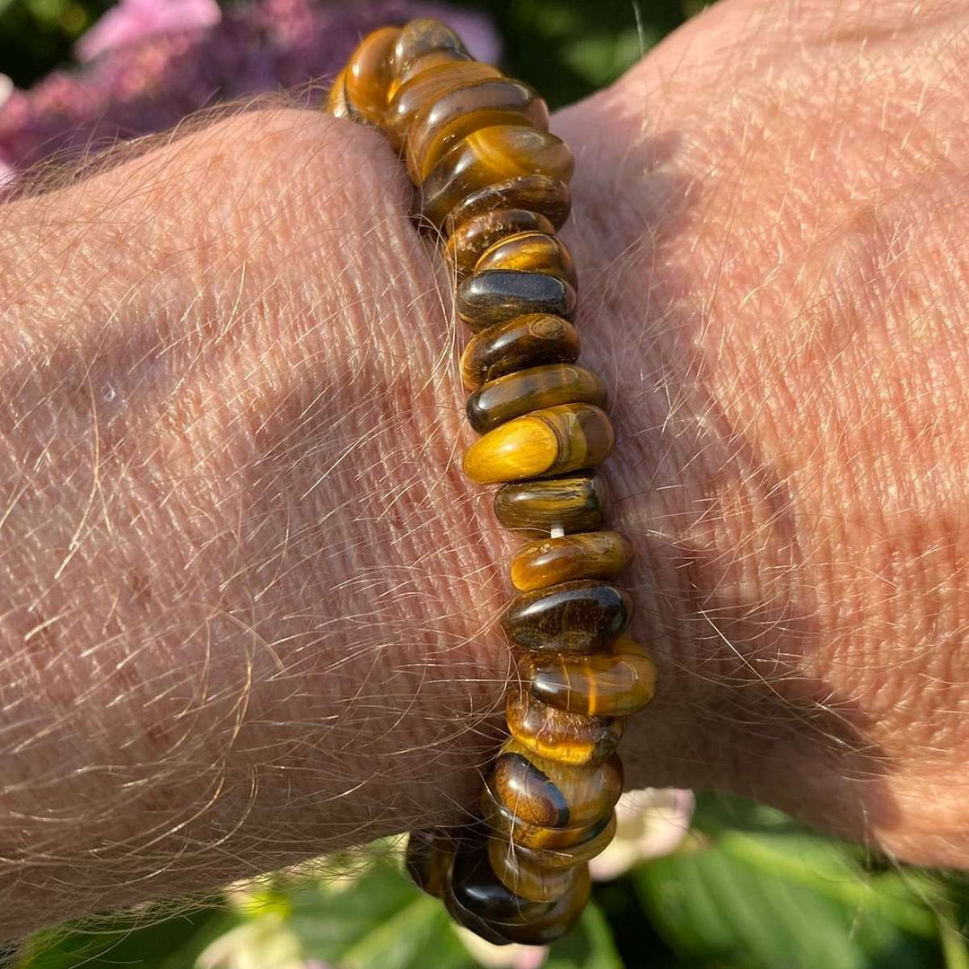 A direct vertical close-up shot of a person's wrist wearing a chunky Tiger Eye crystal bracelet. The large, polished gemstone chips glow with rich golden-yellow bands, chocolate brown tones, and shimmering chatoyant stripes under bright, natural sunlight, contrasted against a soft-focus green garden background.