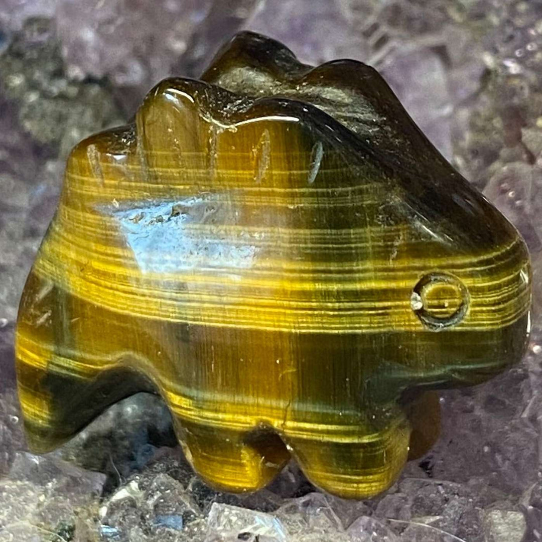 A close-up view of the small, polished Tiger Eye Stegosaurus Carving standing flat. The golden-brown gemstone dinosaur figurine showcases shifting chatoyant stripes and rich honey-coloured bands across its curved back plates, resting beautifully on a raw purple amethyst crystal bed.