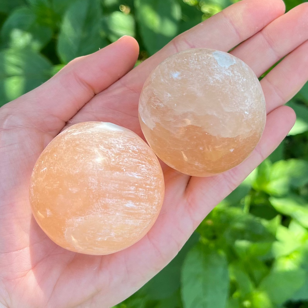 Two polished Peach Selenite crystal balls resting together in an open hand outdoors, displaying their soft internal fractures, luminous chatoyant sheen, and distinct apricot hues under natural sunlight.