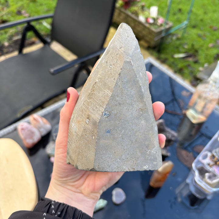 A hand holding the reverse side of the Amethyst freeform block over an outdoor table setting. This view showcases the entirely unpolished, smooth grey natural outer matrix rind and rugged stone backing that highlights its authentic geological origin.