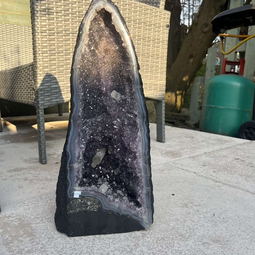 A landscape view focusing on the reverse profile of the upright amethyst geode block. The unpolished, textured green and grey natural outer matrix rind highlights the authentic geological origin and history of this travelling collector&