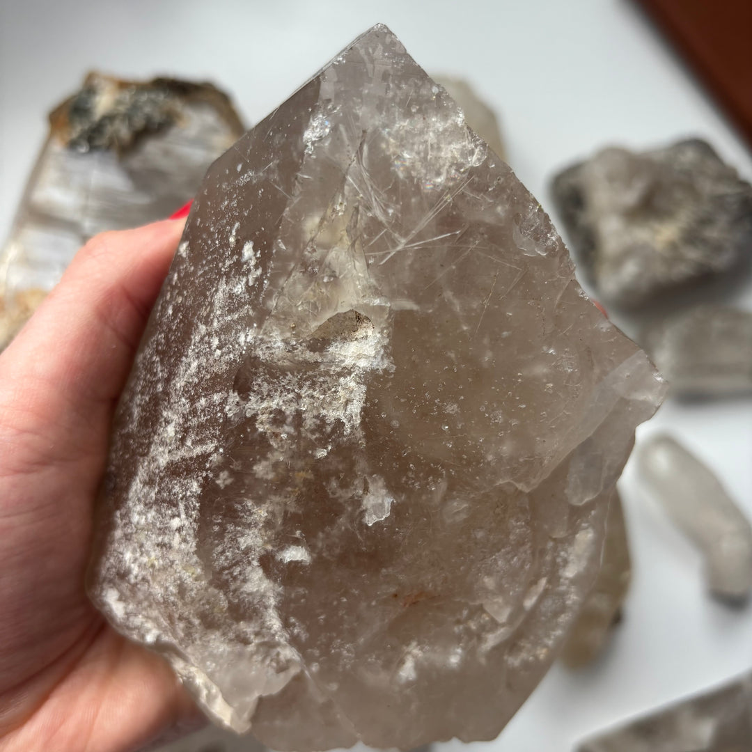 A hand tilting the unpolished clear quartz crystal block to show its large reflective faces, catching the daylight to demonstrate its clean aesthetic, perfect for styling on a prominent shelf or workspace layout.