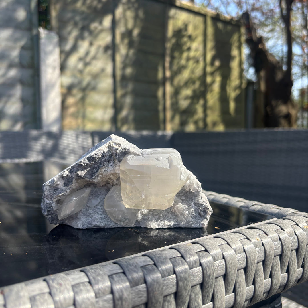 A horizontal display view of the unpolished grey chalcedony and calcite cluster sitting on textured garden furniture, highlighting the synergistic magic and earthy contours of this 9x8cm geological statement piece.
