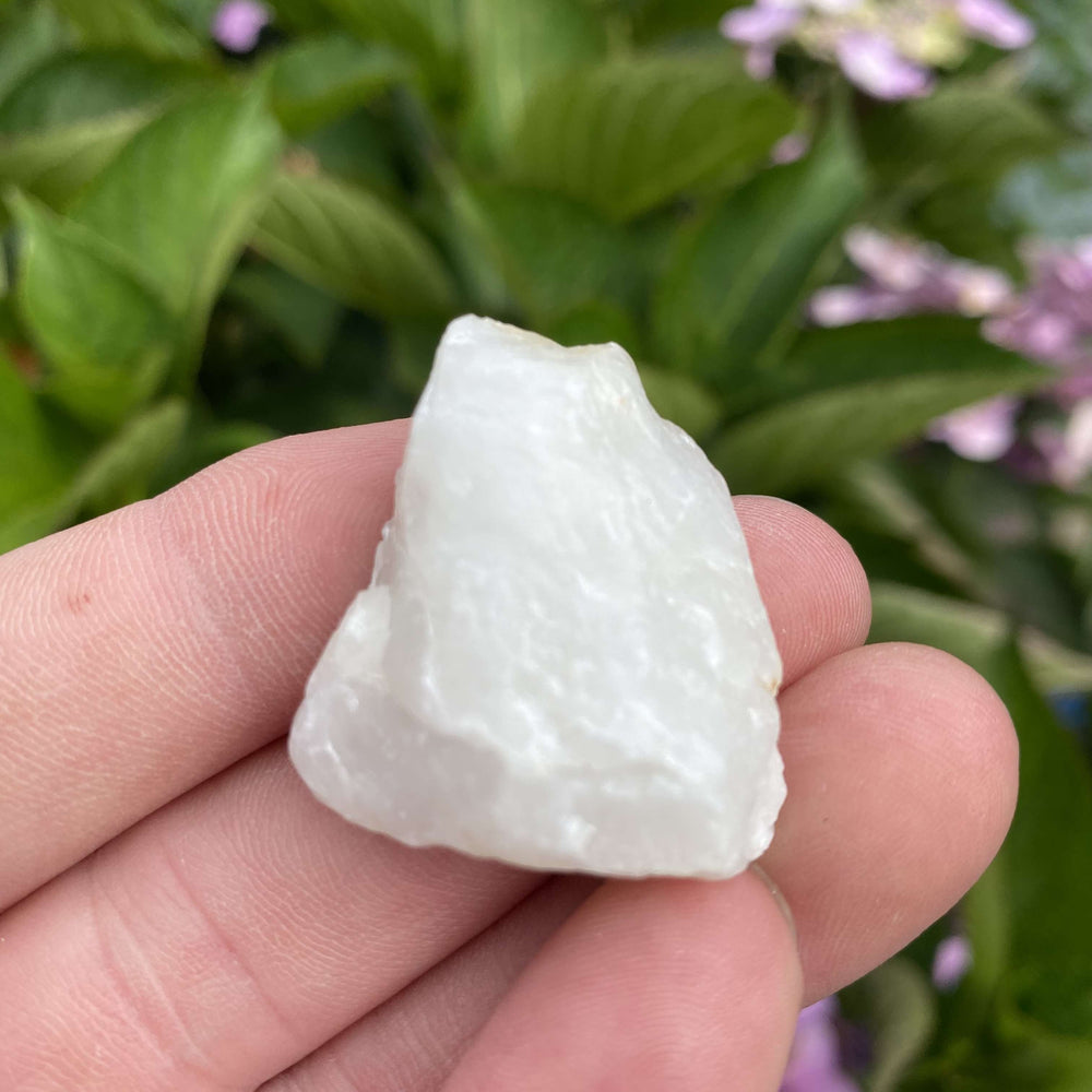 A hand holding a rough, unpolished milky white quartz stone outdoors, displaying its natural frosty peaks and geometric mineral ridges.
