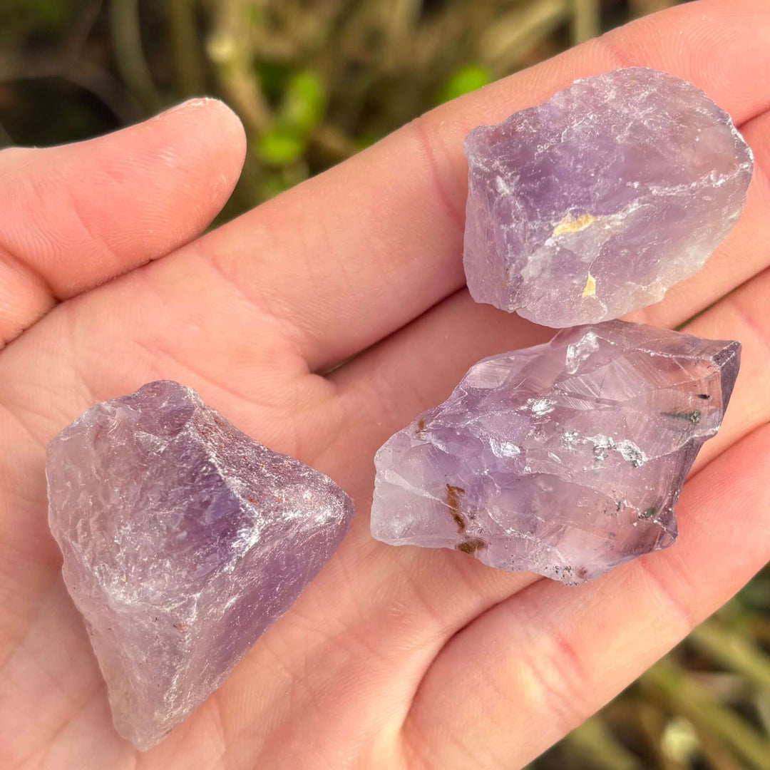 A tight close-up view of three individual raw Amethyst chunks cradled across an open palm outdoors. This perspective highlights the rough texture, unpolished crystal points, and unique crystalline shapes of each natural pocket stone under bright daylight.