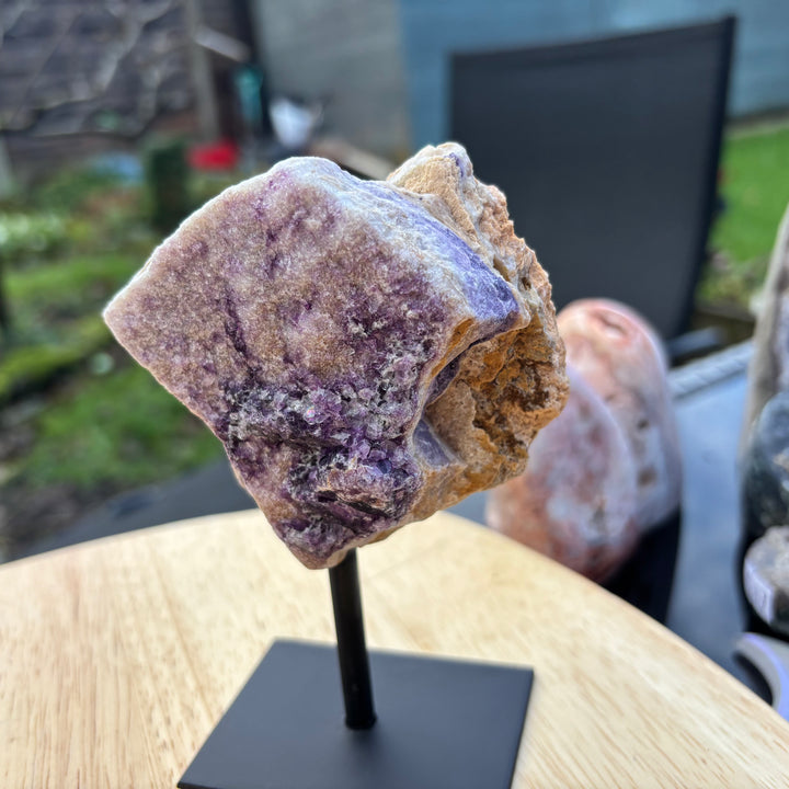 A slightly angled view of the purple Raw Fluorite display piece resting on its custom black metal stand outdoors. This unpolished, light-coloured matrix stone acts as a brilliant visual anchor to quiet a busy mind, bring deep calm to everyday emotions, and align perfectly with the Crown Chakra.