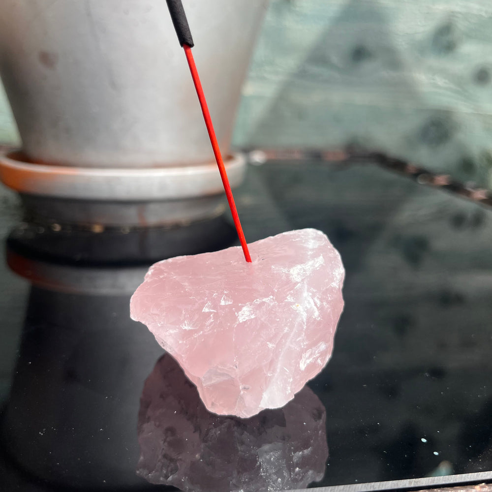 A vertical close-up shot focusing tightly on the unpolished Rose Quartz incense burner block with an incense stick inserted into its pre-drilled slot. The soft daylight accentuates the delicate pink colour variations and translucent quartz texture, ideal for balancing the Heart Chakra.
