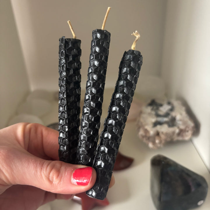A tight close-up view focusing on the detailed geometric honeycomb pattern of three black rolled beeswax ritual candles. Held firmly in a hand with pink polish, this shot highlights the raw organic texture built for clearing away heavy energy during a cosy evening shadow work routine.