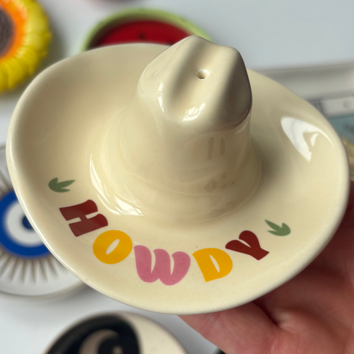 A close-up view from a slightly elevated angle focusing on the smooth glazed finish and colourful "HOWDY" lettering on the front brim of the cowboy hat burner. The holder sits cradled in a hand to highlight its neat 10cm proportions and playful Western style under bright, natural daylight.
