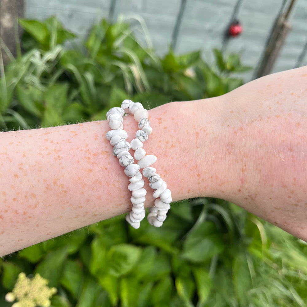 A wider shot of a wrist presenting two stacked white howlite gemstone chip bracelets outdoors, illustrating the stretchy 18cm flexible sizing scale and comfy everyday fit against sunlit garden greenery.