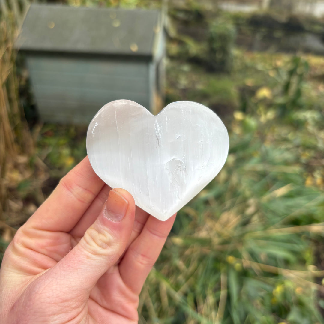 A hand displaying a single white selenite puffy heart crystal token upright against a soft outdoor greenery background, showcasing its symmetrical carved silhouette, perfect for sliding into a coat pocket or purse.
