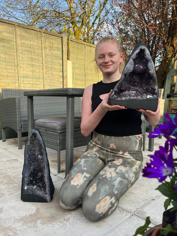 A person kneeling outdoors on a patio holds a large polished black geode specimen, with a second geode on the ground nearby. The background includes patio furniture, a wooden fence, trees, and purple flowers. 