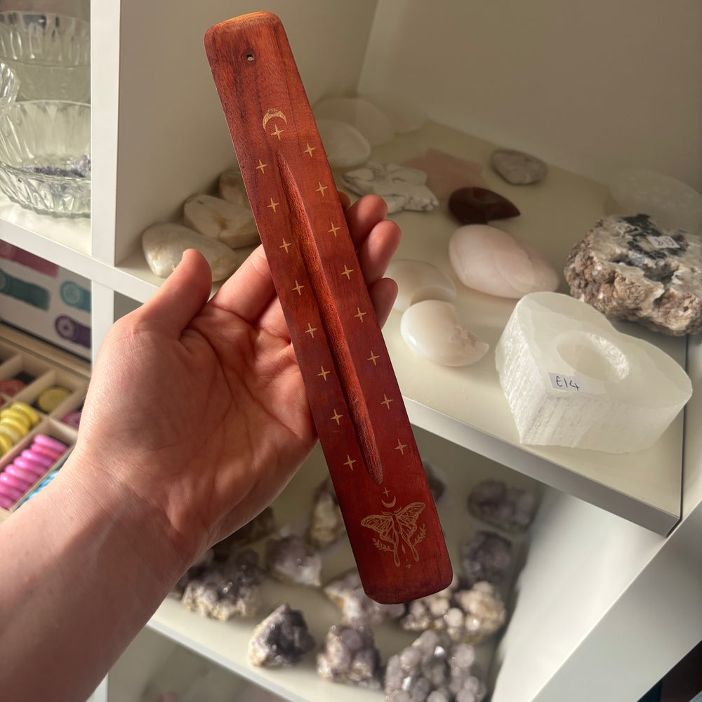An alternative high-angle indoor shot of the mystical moth incense tray held in a hand to demonstrate its physical 25cm length. The background displays white retail display cubbies filled with polished stones and raw white crystals under soft lighting.