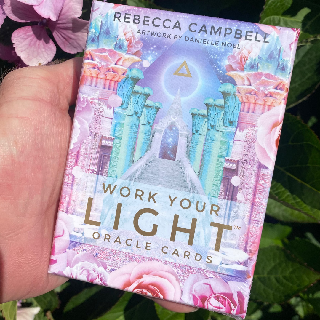 A direct vertical close-up shot of a person's hand holding the Work Your Light Oracle Cards box outdoors. The packaging features a vibrant, pastel pink and blue fantasy illustration of a glowing cosmic temple by Danielle Noel with gold lettering, set against a soft-focus background of green leaves and pink flowers.