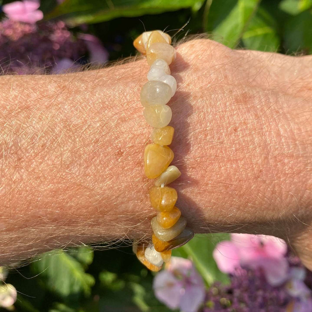 A side-angle outdoor view of the stretchy crystal bracelet worn on a wrist. The individual, sunny yellow jade chips catch the light, showing off their natural, translucent properties against a textured background of green garden leaves and soft pink blossoms.
