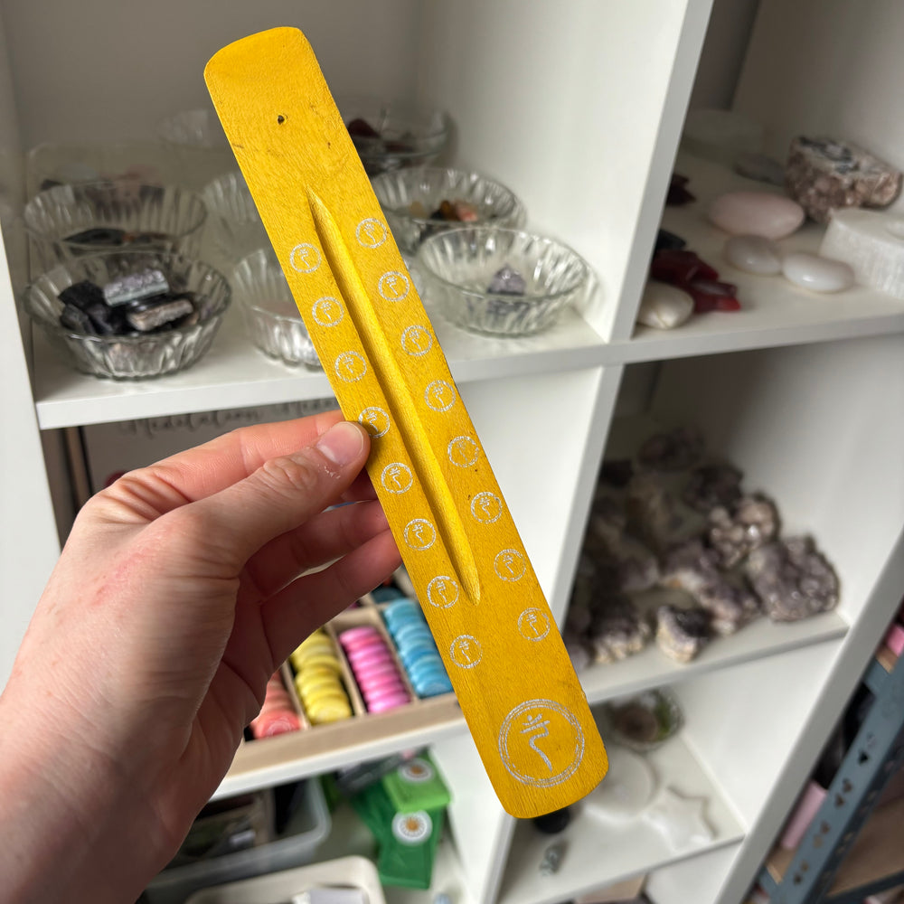An eye-level shot showing the yellow wooden incense holder held flat in a hand to highlight its rustic warmth and long central ash-catching groove. The sleek, slim profile is displayed before a shop background, showing how it creates a cosy everyday vibe on any side table.