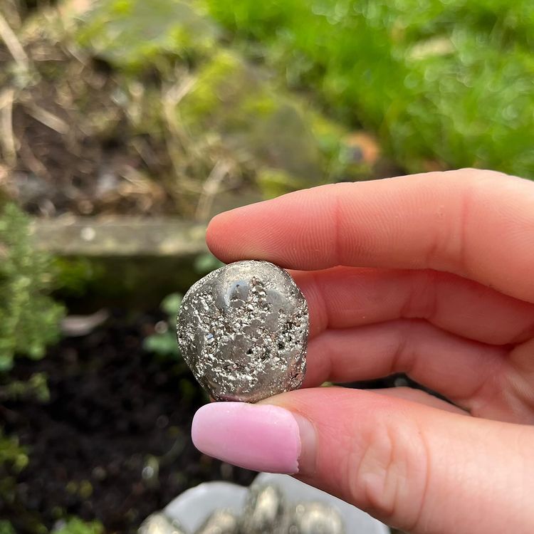 A hand holding a Pyrite Tumble Stone in a bright sun lit garden