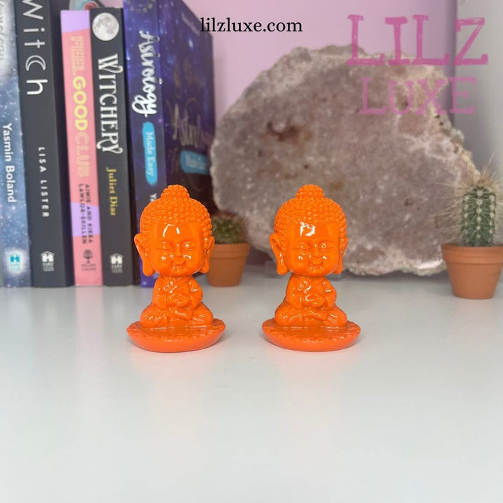 Two orange incense burners shaped like baby Buddhas, placed on a shelf with books and a decorative stone in the background.
