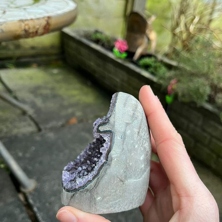 A side profile shot of the amethyst cut base cluster held in a hand, displaying the smooth, light gray polished agate rind wrapping around the crystal point.