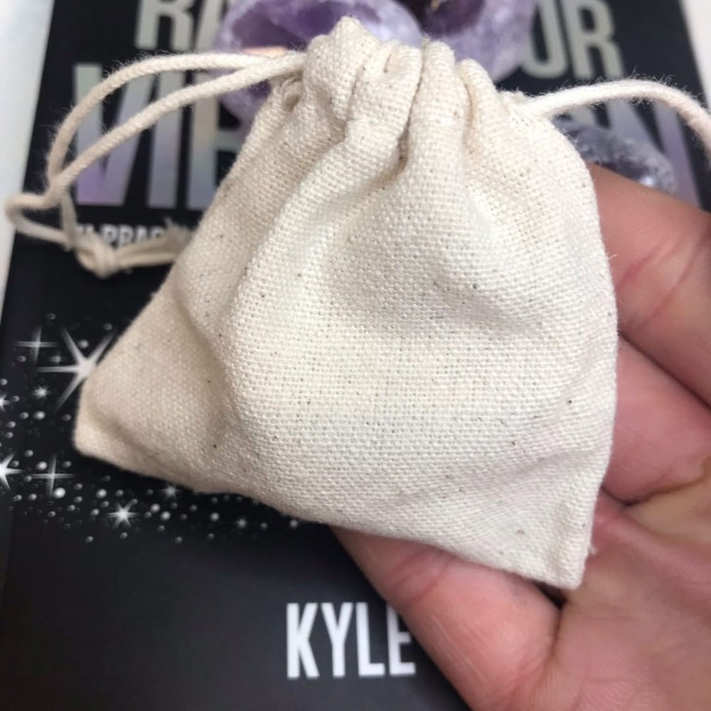 Close-up of a person holding a small, natural off-white calico or cotton drawstring jewellery pouch, with a black book cover partially visible in the background.
