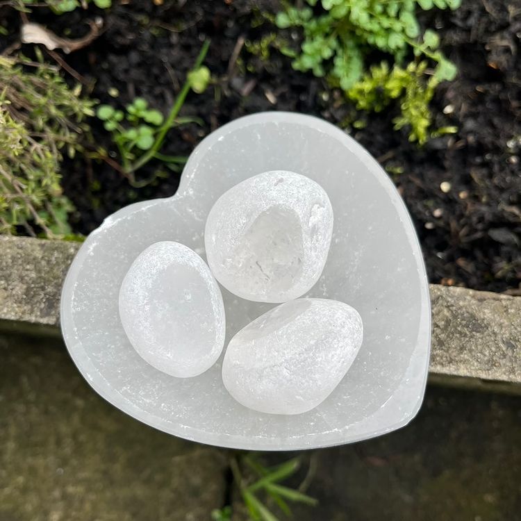 A top-down outdoor view of three smooth, polished translucent clear quartz tumble stones arranged inside a carved white selenite heart-shaped crystal bowl on a stone surface