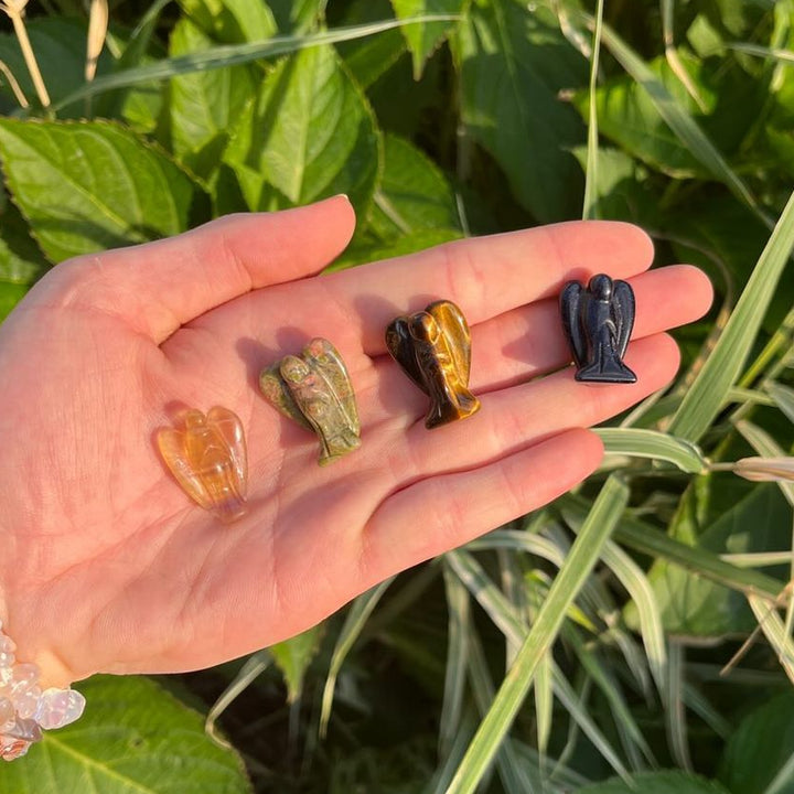 A hand holding four different colored crystal carvings of angels, with a background of green plant leaves.