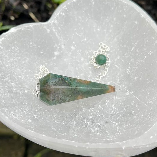 A heart-shaped green moss agate crystal pendulum with a silver chain, placed on a heart-shaped stone.