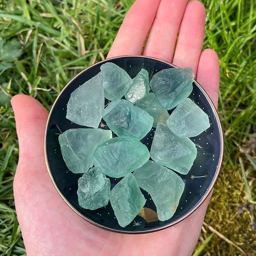 A collection of mini raw Green Fluorite crystals held in an open hand, showcasing their rough unpolished edges, unique geometric growth layers, and soft minty green colours against a black backdrop.