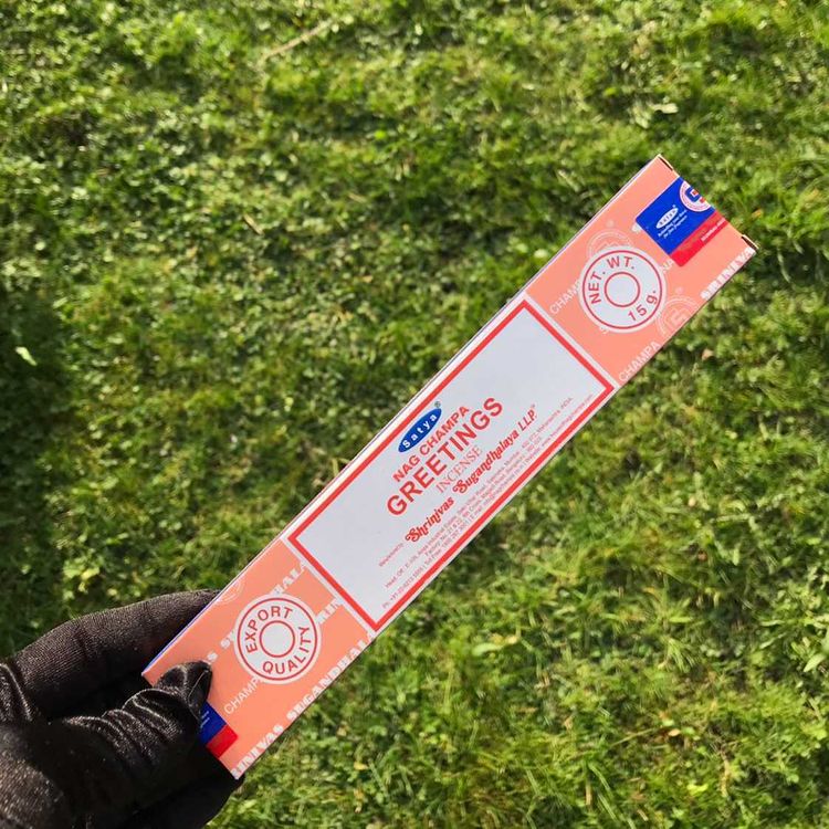 A pack of Satya Greetings incense sticks held in a person's hand, with the packaging displaying the product name and branding against a grassy background.