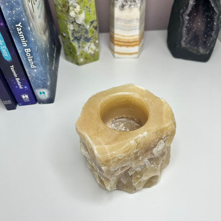 A beige honey calcite tea light holder placed on a shelf with various crystal decorations and books in the background.