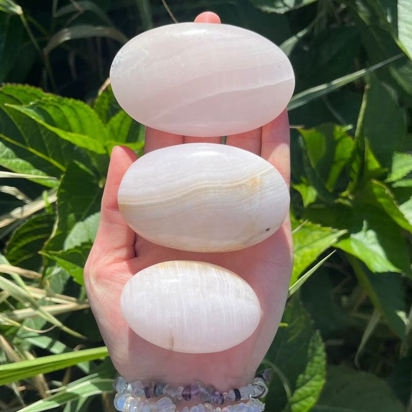 Three smooth, polished Mangano Calcite Palm Stones with subtle natural banding are held in a person's hand against a background of green outdoor foliage.