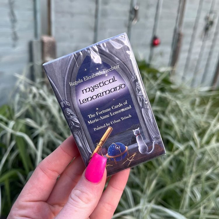 A hand holding a pack of Mystical Lenormand Mini Oracle Cards by Urban Trosch in a brightly sun lit garden