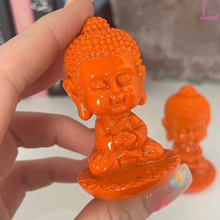 A hand holds a small Orange Baby Buddha Stick Incense Burner with a meditative pose, with another similar figurine blurred in the background.
