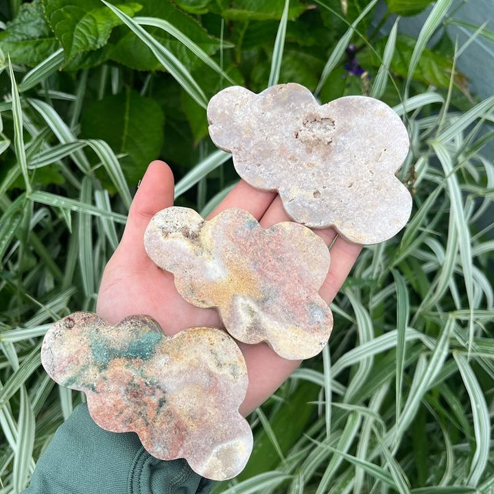Three pink amethyst crystal figures with varying cloud-like shapes held in a hand, with green plant background.