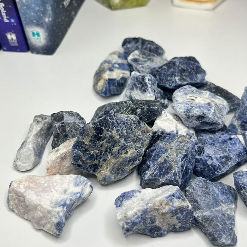 A collection of raw sodalite crystals displayed on a white surface, with books and a candle in the background.