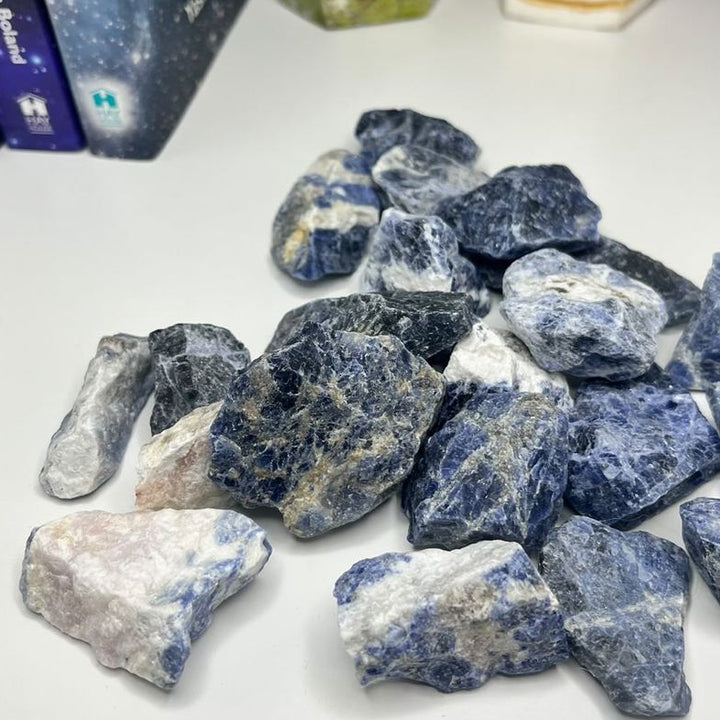 A collection of raw sodalite crystals displayed on a white surface, with books and a candle in the background.