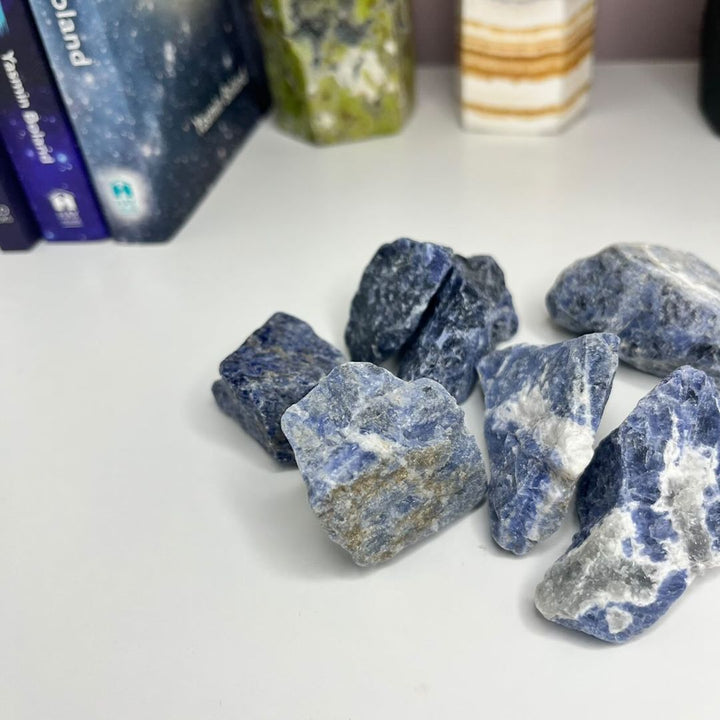 A collection of raw sodalite crystals displayed on a white surface, with books and a candle in the background.