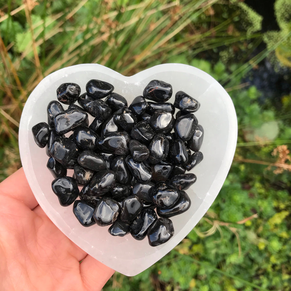 A hand holding a collection of tiny black onyx tumble stones shaped like a heart, with a blurred green background.