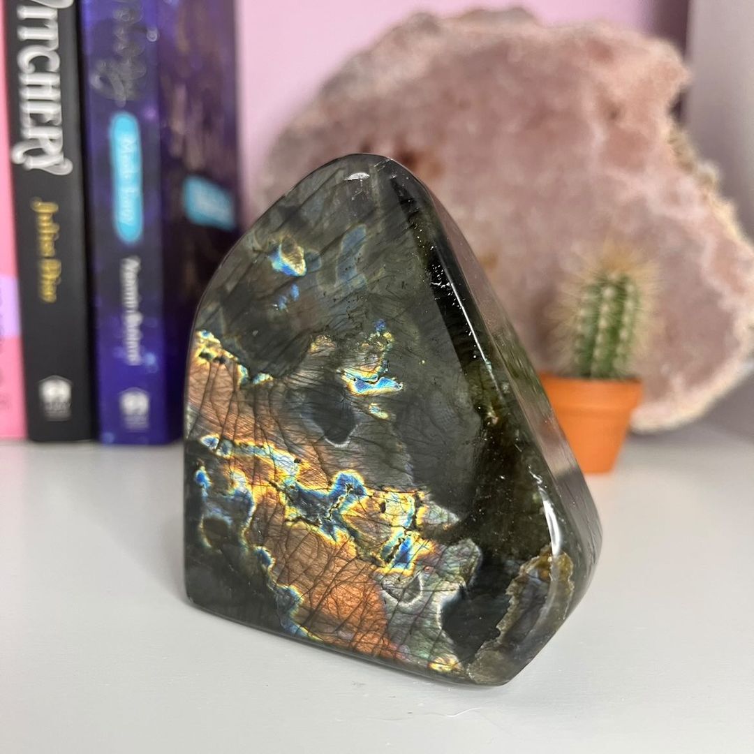 An A Grade Labradorite polished freeform crystal statement piece standing on its carved base, displaying rich cosmic flashes of fiery gold and peacock blue, styled indoors next to a tiny potted cactus and spiritual books for Third Eye alignment.